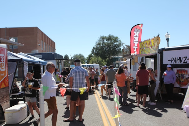 A lively outdoor market scene with people walking and conversing between food trucks parked on either side of a street. Brightly colored banners and flags, like 'Kettle Corn', are visible and add to the festive atmosphere. The setting is under clear blue sky with a distant view of trees and buildings.