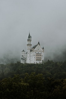 A grand, fairytale-like castle stands majestically atop a forested hill. Its towers and spires pierce through the mist and clouds that cover the scene, creating a mystical atmosphere.