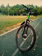 A bicycle is parked on a paved track in a park-like setting. The background shows a grassy field with a soccer goal and dense green trees under a clear sky.