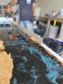 A person appears to be working with a mold, possibly creating a resin and wood table or countertop. The image prominently features blue swirling resin being poured and shaped within a wooden frame. Various tools and equipment are present in the background, indicating a workshop setting.