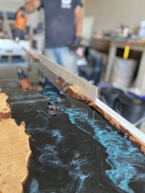 A person appears to be working with a mold, possibly creating a resin and wood table or countertop. The image prominently features blue swirling resin being poured and shaped within a wooden frame. Various tools and equipment are present in the background, indicating a workshop setting.