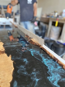 A person appears to be working with a mold, possibly creating a resin and wood table or countertop. The image prominently features blue swirling resin being poured and shaped within a wooden frame. Various tools and equipment are present in the background, indicating a workshop setting.
