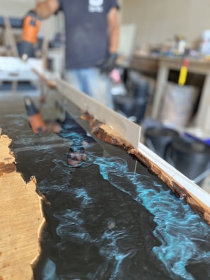 A person appears to be working with a mold, possibly creating a resin and wood table or countertop. The image prominently features blue swirling resin being poured and shaped within a wooden frame. Various tools and equipment are present in the background, indicating a workshop setting.