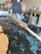 A person appears to be working with a mold, possibly creating a resin and wood table or countertop. The image prominently features blue swirling resin being poured and shaped within a wooden frame. Various tools and equipment are present in the background, indicating a workshop setting.