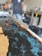 A person appears to be working with a mold, possibly creating a resin and wood table or countertop. The image prominently features blue swirling resin being poured and shaped within a wooden frame. Various tools and equipment are present in the background, indicating a workshop setting.