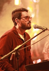 A man is seen performing music, positioned at a microphone. He is wearing glasses and headphones and dressed in a brown shirt. The warm lighting creates a soft, intimate atmosphere with a mic stand and keyboard visible.