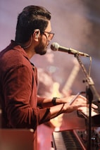 A musician plays a keyboard while singing into a microphone. He wears glasses and a brownish shirt, and the stage is lit with a warm, ambient glow. In the background, a fellow band member and a guitar can be seen, slightly out of focus, contributing to a dynamic performance atmosphere.
