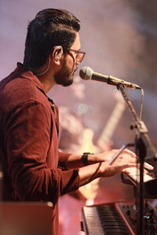 A musician plays a keyboard while singing into a microphone. He wears glasses and a brownish shirt, and the stage is lit with a warm, ambient glow. In the background, a fellow band member and a guitar can be seen, slightly out of focus, contributing to a dynamic performance atmosphere.
