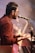 A musician plays a keyboard while singing into a microphone. He wears glasses and a brownish shirt, and the stage is lit with a warm, ambient glow. In the background, a fellow band member and a guitar can be seen, slightly out of focus, contributing to a dynamic performance atmosphere.