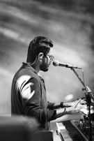 A person wearing glasses and a dark shirt is playing a keyboard while singing into a microphone. The scene is captured in black and white, highlighting the contrast and shadows. The focus is on the musician, and the surroundings are blurred, creating an intimate and artistic atmosphere.