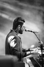 A person wearing glasses and a dark shirt is playing a keyboard while singing into a microphone. The scene is captured in black and white, highlighting the contrast and shadows. The focus is on the musician, and the surroundings are blurred, creating an intimate and artistic atmosphere.
