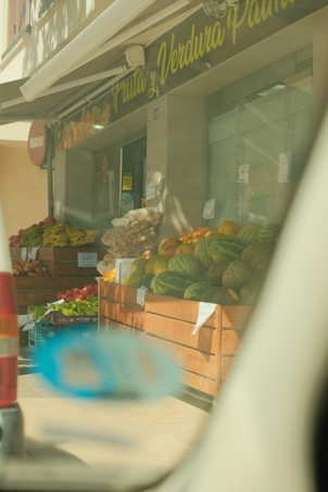 A market stall with a variety of fruits on display, including watermelons, bananas, and other colorful produce. The stand is outside a store with a sign in Spanish, and the view is partially obscured by a car window.