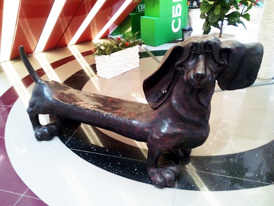 A large sculpture of a dachshund with exaggerated features, including long ears and an elongated body. The sculpture is situated indoors on a tiled floor with reflective surfaces. Plants and modern decorative elements are visible in the background.