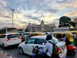 A busy city street scene with a diverse mix of vehicles including cars, motorcycles, and auto-rickshaws in traffic. In the background, an impressive government building with ornate architectural details is visible against a partly cloudy sky at sunset. A large tree is also visible in the scene.