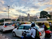 A busy city street scene with a diverse mix of vehicles including cars, motorcycles, and auto-rickshaws in traffic. In the background, an impressive government building with ornate architectural details is visible against a partly cloudy sky at sunset. A large tree is also visible in the scene.