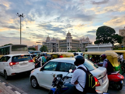 A busy city street scene with a diverse mix of vehicles including cars, motorcycles, and auto-rickshaws in traffic. In the background, an impressive government building with ornate architectural details is visible against a partly cloudy sky at sunset. A large tree is also visible in the scene.