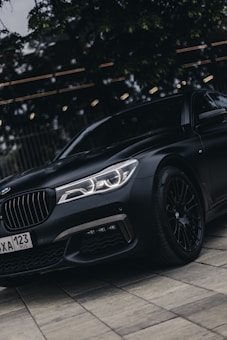 A sleek, black luxury sedan is parked on a tiled surface, subtly reflecting ambient light. The vehicle has a visible license plate, precision-engineered headlights, and a polished finish. The surrounding area includes some trees and possibly artificial lighting, offering a reflective environment.