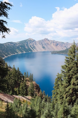 A serene landscape depicting a deep blue lake surrounded by lush green pine forests and towering mountain peaks. Fluffy white clouds float across a bright blue sky, enhancing the tranquility and natural beauty of the scene.