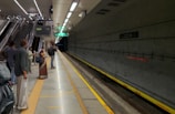 A group of people waits on a clean and well-lit subway platform, visible are tracks and various signage displaying directions and train information. The platform features modern design elements with concrete walls and an illuminated ceiling. People are standing near the edge, looking down the tunnel as they anticipate an approaching train.