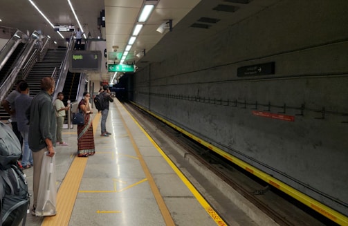 A group of people waits on a clean and well-lit subway platform, visible are tracks and various signage displaying directions and train information. The platform features modern design elements with concrete walls and an illuminated ceiling. People are standing near the edge, looking down the tunnel as they anticipate an approaching train.