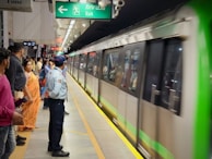 A busy metro station platform with a green and silver train arriving. Passengers are standing in orderly lines along the platform, including a security officer in a light blue uniform. An exit sign is visible above, and several people are seen waiting patiently.