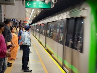 A busy metro station platform with a green and silver train arriving. Passengers are standing in orderly lines along the platform, including a security officer in a light blue uniform. An exit sign is visible above, and several people are seen waiting patiently.