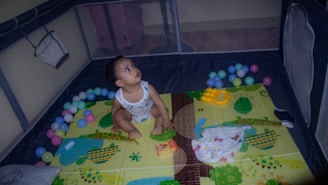 An infant is seated in a colorful playpen surrounded by multicolored plastic balls. The play area has a vibrant mat with playful designs including trees and animals. The baby is looking upwards, wearing a sleeveless outfit. Various toys and a diaper are scattered around.