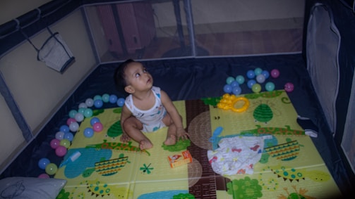 An infant is seated in a colorful playpen surrounded by multicolored plastic balls. The play area has a vibrant mat with playful designs including trees and animals. The baby is looking upwards, wearing a sleeveless outfit. Various toys and a diaper are scattered around.