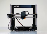 A 3D printer with a black frame and a visible extruder head equipped with wiring and motors. It rests on a clean, white surface with metal rods and a flat print bed for creating three-dimensional objects.