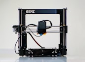 A 3D printer with a black frame and a visible extruder head equipped with wiring and motors. It rests on a clean, white surface with metal rods and a flat print bed for creating three-dimensional objects.
