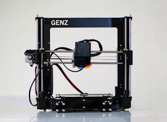 A 3D printer with a black frame and a visible extruder head equipped with wiring and motors. It rests on a clean, white surface with metal rods and a flat print bed for creating three-dimensional objects.