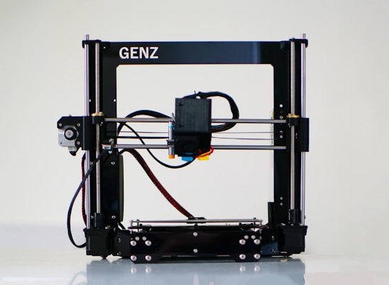 A 3D printer with a black frame and a visible extruder head equipped with wiring and motors. It rests on a clean, white surface with metal rods and a flat print bed for creating three-dimensional objects.