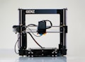 A 3D printer with a black frame and a visible extruder head equipped with wiring and motors. It rests on a clean, white surface with metal rods and a flat print bed for creating three-dimensional objects.