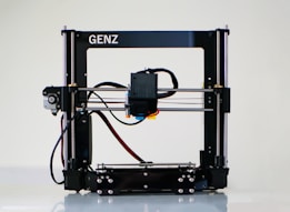A 3D printer with a black frame and a visible extruder head equipped with wiring and motors. It rests on a clean, white surface with metal rods and a flat print bed for creating three-dimensional objects.