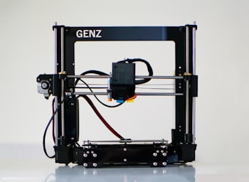 A 3D printer with a black frame and a visible extruder head equipped with wiring and motors. It rests on a clean, white surface with metal rods and a flat print bed for creating three-dimensional objects.