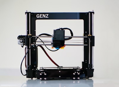 A 3D printer with a black frame and a visible extruder head equipped with wiring and motors. It rests on a clean, white surface with metal rods and a flat print bed for creating three-dimensional objects.