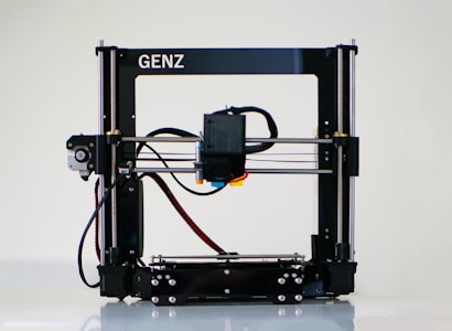 A 3D printer with a black frame and a visible extruder head equipped with wiring and motors. It rests on a clean, white surface with metal rods and a flat print bed for creating three-dimensional objects.