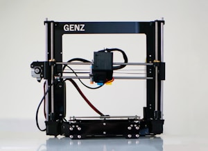 A 3D printer with a black frame and a visible extruder head equipped with wiring and motors. It rests on a clean, white surface with metal rods and a flat print bed for creating three-dimensional objects.