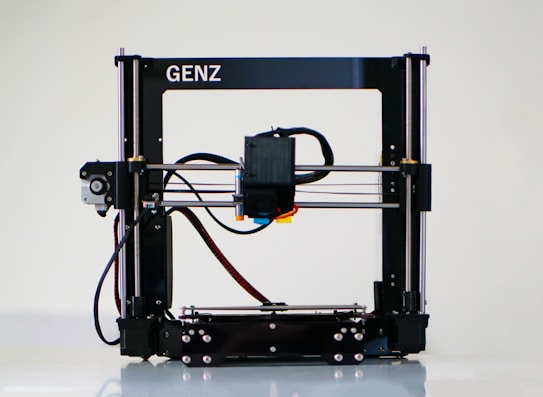 A 3D printer with a black frame and a visible extruder head equipped with wiring and motors. It rests on a clean, white surface with metal rods and a flat print bed for creating three-dimensional objects.