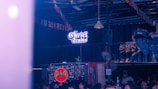 A dimly lit indoor space with a sign reading 'The Cartel Studio' illuminated in neon. The walls are decorated with various graffiti and stickers, including a prominent Bacardi logo. The ceiling is adorned with colorful triangular flags. Several people are noticeable, suggesting a social gathering.
