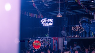A dimly lit indoor space with a sign reading 'The Cartel Studio' illuminated in neon. The walls are decorated with various graffiti and stickers, including a prominent Bacardi logo. The ceiling is adorned with colorful triangular flags. Several people are noticeable, suggesting a social gathering.