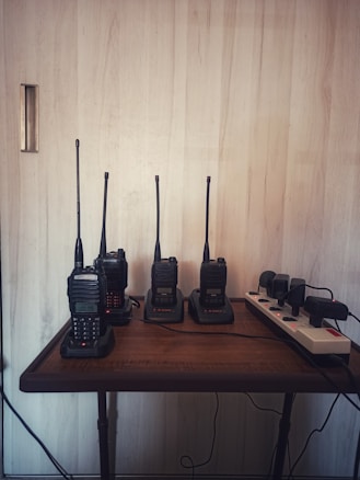 Four walkie-talkies are neatly placed on a wooden table, each positioned in its own charging dock. A power strip with multiple outlets is placed to the right, with cables connected to each device. The background consists of a light-colored wooden wall, creating a minimalist and organized setting.