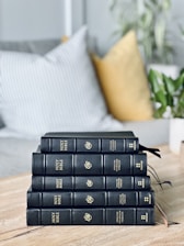 A stack of five black leather-bound books titled 'Holy Bible' with gold lettering is neatly arranged on a wooden table. In the background, cushions in muted and patterned colors are placed on a sofa, and a potted plant adds a touch of greenery.
