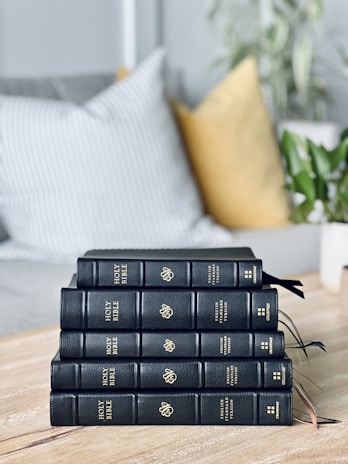 A stack of five black leather-bound books titled 'Holy Bible' with gold lettering is neatly arranged on a wooden table. In the background, cushions in muted and patterned colors are placed on a sofa, and a potted plant adds a touch of greenery.