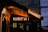 A dimly lit exterior of a bookstore with glowing neon signs in Cyrillic script, possibly Russian, indicating the presence of books. The building has a rustic facade with a wooden awning, and street signs are visible nearby, suggesting an urban setting.