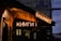 A dimly lit exterior of a bookstore with glowing neon signs in Cyrillic script, possibly Russian, indicating the presence of books. The building has a rustic facade with a wooden awning, and street signs are visible nearby, suggesting an urban setting.