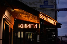A dimly lit exterior of a bookstore with glowing neon signs in Cyrillic script, possibly Russian, indicating the presence of books. The building has a rustic facade with a wooden awning, and street signs are visible nearby, suggesting an urban setting.