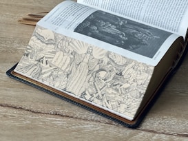 A large, open book is laid on a wooden surface. The pages feature black and white illustrations with religious themes, showcasing various biblical scenes including figures in robes and symbolic imagery. The text is in English and appears to be part of the Old Testament.