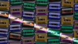 Various cassette tapes in different colors are scattered, showcasing vibrant designs and labels. The tapes are arranged in an overlapping pattern, creating a retro and nostalgic feel.