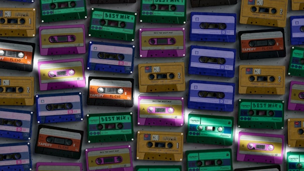 Various cassette tapes in different colors are scattered, showcasing vibrant designs and labels. The tapes are arranged in an overlapping pattern, creating a retro and nostalgic feel.